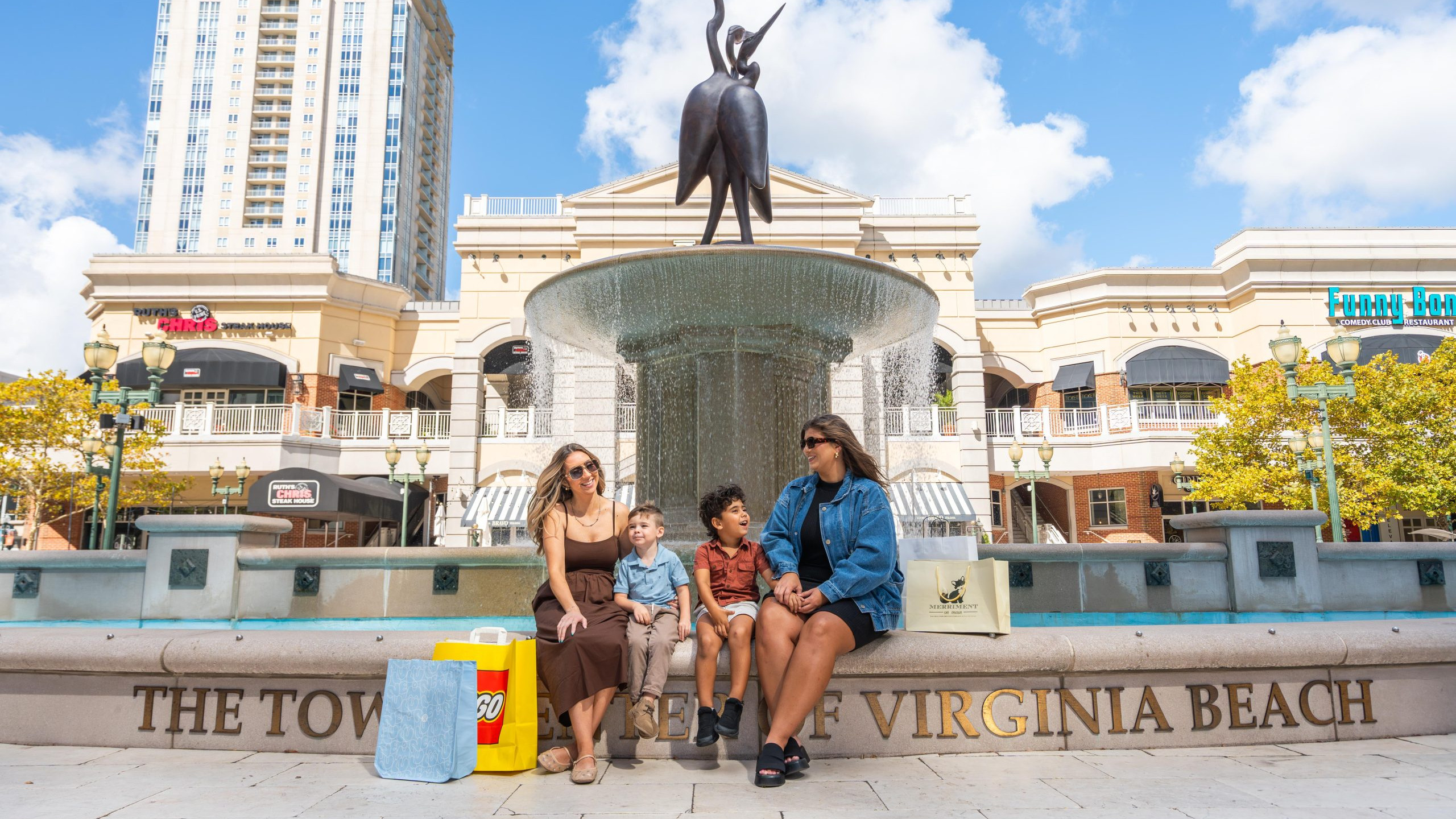 2 moms with their 2 sons sitting at the fountain plaza with shopping bags